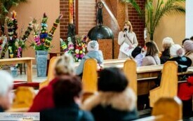 Ludzie siedzący w ławkach kościelnych słuchający przem&oacute;wienia kobiety w płaszczu