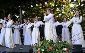 Dziewczyny w białych sukniach i wiankach na głowach śpiewają na scenie