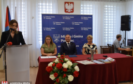 Kobieta przemawia na m&oacute;wnicy, troje ludzi siedzi przy stole