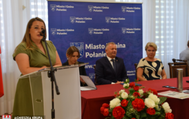 Kobieta przemawia na m&oacute;wnicy, troje ludzi siedzi przy stole