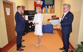 po lewej stronie na zdjęciu burmistrz Jacek Nowak, kt&oacute;ry gratuluje nauczycielce, kt&oacute;ra znajduje się pomiędzy burmistrzem a przewodniczącym Rady Miejskiej Stanisławem Lolo kt&oacute;ry znajduje się po prawej stronie i kt&oacute;ry trzyma bukiet z czerwonych r&oacute;ż