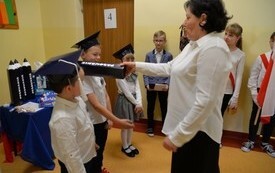 na zdjęciu znajduje się 7 os&oacute;b, pani dyrektor szkoły w Zrębinie dokonuje pasowania uczni&oacute;w klas pierwszych, w oddali widać stolik nakryty niebieskim obrusem na kt&oacute;rym są oł&oacute;wki r&oacute;żnej wielkości