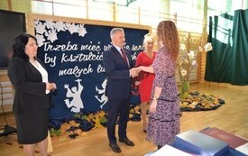 na zdjęciu po lewej stronie znajduje się dyrektor szkoły w Ruszczy ubrana w czarny żakiet oraz białą bluzkę, kolejno burmistrz miasta i gminy Połaniec Jacek Nowak kt&oacute;ry gratuluje nauczycielce, kt&oacute;ra stoi przed nim w fioletowej sukience w kwiatki oraz po p
