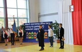 grupa trzech uczni&oacute;w kt&oacute;rzy przemawiają do uczni&oacute;w, po lewej poczet sztandarowy a za nim dwie nauczycielki