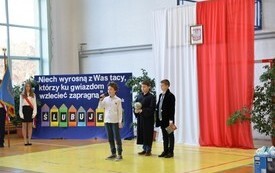 na zdjęciu grupa trzech uczni&oacute;w chłopc&oacute;w, gdzie jeden z nich przemawia do publiczności, w oddali po lewej stronie poczet sztandarowy