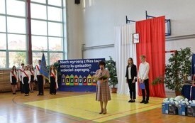 pani dyrektor wygłasza przem&oacute;wienie do pierwszoklasist&oacute;w, a za nią dw&oacute;ch chłopc&oacute;w, natomiast po lewej stronie stoi poczet sztandarowy
