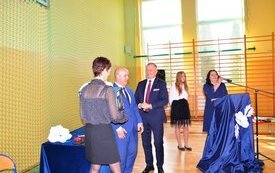 Burmistrz, radny wręczają prezent wychowawcy w tle dyrektor i uczennica