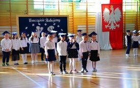 Grupka dzieci podczas wystąpienia na sali w tle godło Polski, tablica do kosza