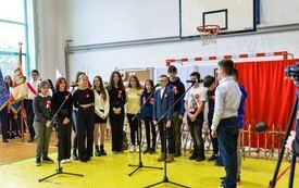 grupa uczni&oacute;w kt&oacute;rzy śpiewają, uczniowie stają w p&oacute;łkolu, przy mikrofonach jest 13 os&oacute;b, w oddali po lewej strony poczet sztandarowy