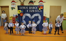 na zdjęciu dzieci z przedszkola dziewczynki białe bluzki i sukienki w kwiatki, zaś chłopcy ciemne spodnie, białe koszule i szelki, po bokach nauczycielki