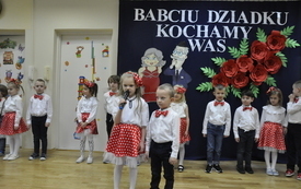 Na zdjęciu widoczny jest chłopiec z dziewczynką trzymający mikrofon i recytujący wiersz. W tle widać pozostałe dzieci biorące udział w przedstawieniu